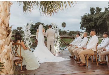 Wedding ceremony on the wooden deck at the water's edge, guests seated under palm trees beside the lagoon